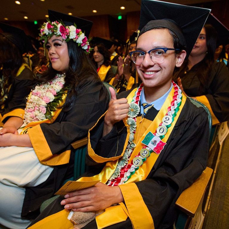 Graduates in caps and gowns at a ceremony, one giving a thumbs-up and wearing a lei.